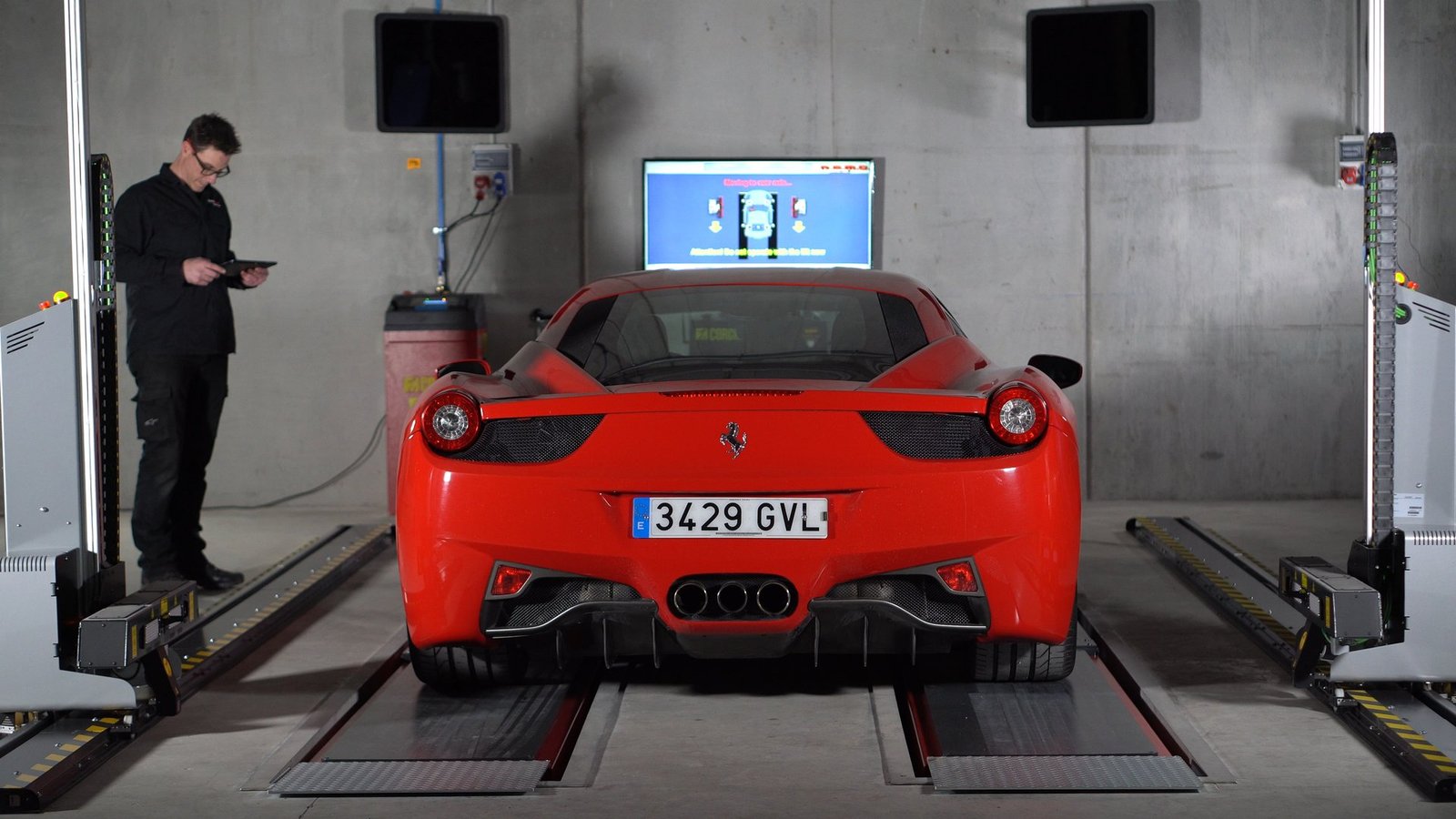 Ferrari rojo en máquina de alineación de ruedas en taller. Un técnico con gafas revisa datos en una tablet junto al coche.